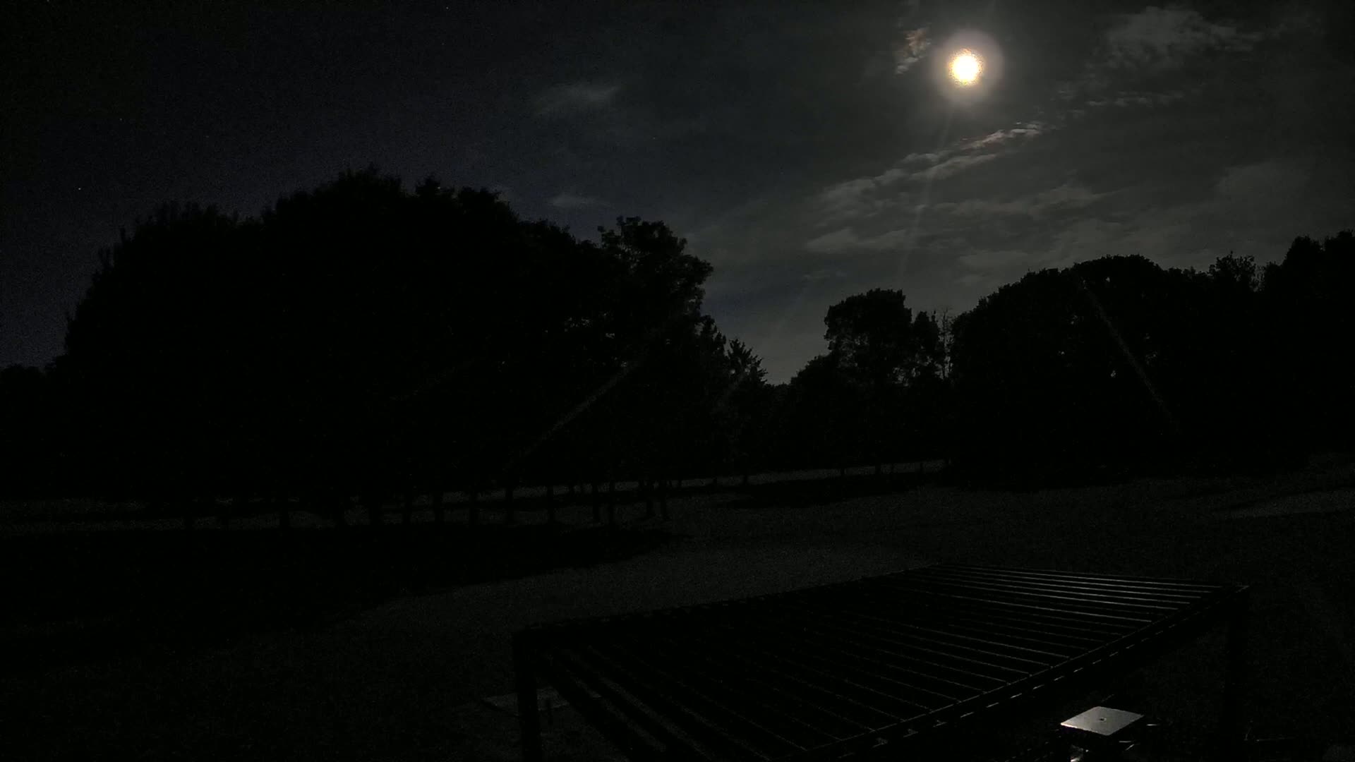 A bright moon shines through partly cloudy night sky over a dark, silhouetted landscape of trees and a metal grate in the foreground.