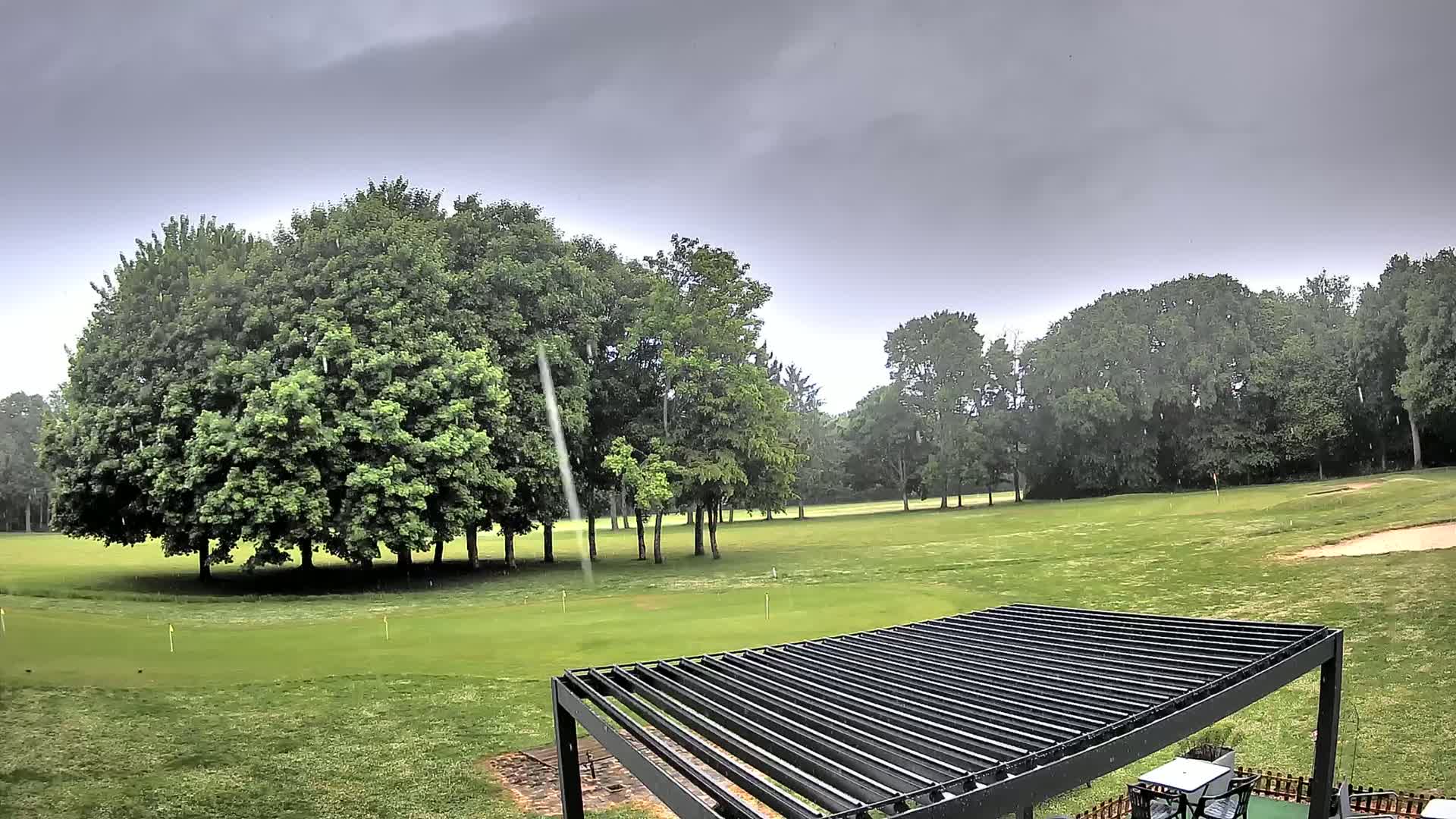 Rain falls on a green golf course surrounded by lush, green trees under a gray sky.