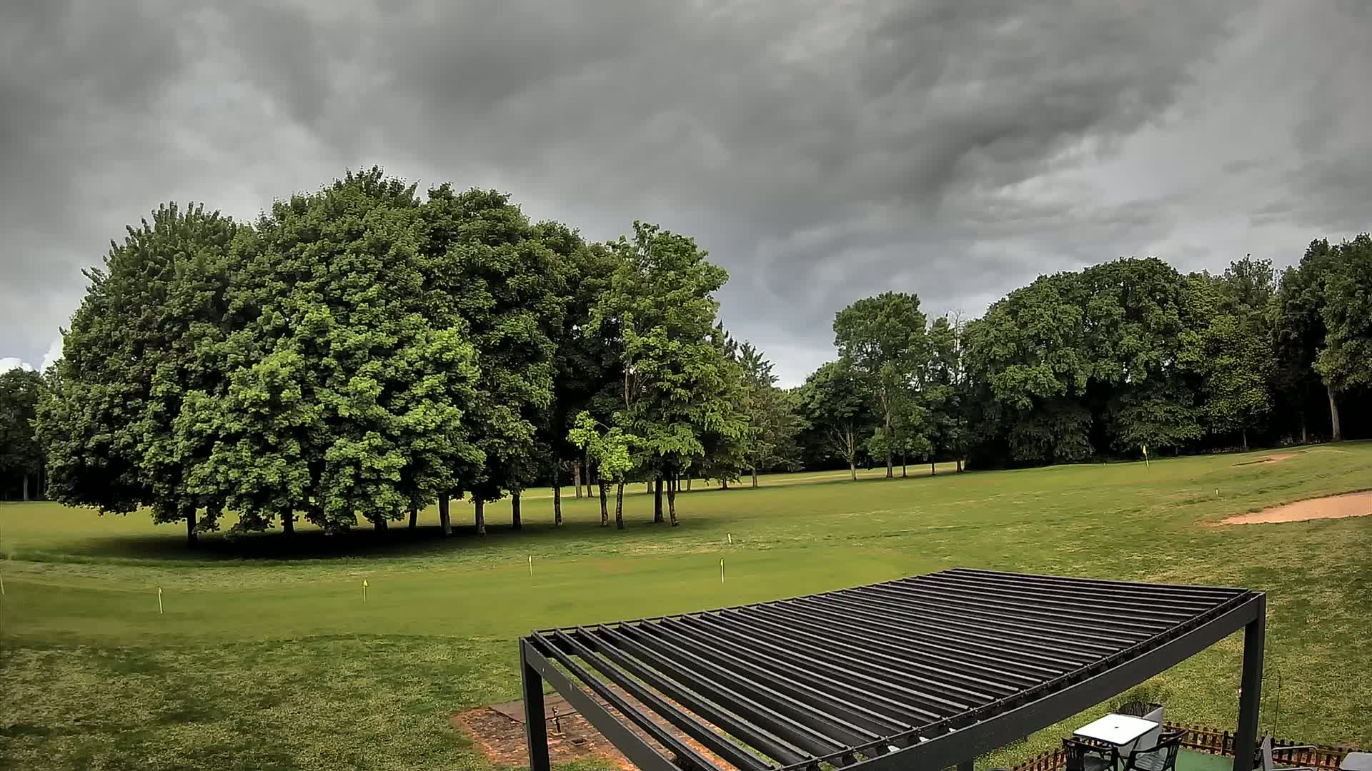 A dark, cloudy sky hangs over a green golf course with several leafy trees and a black pergola in the foreground.