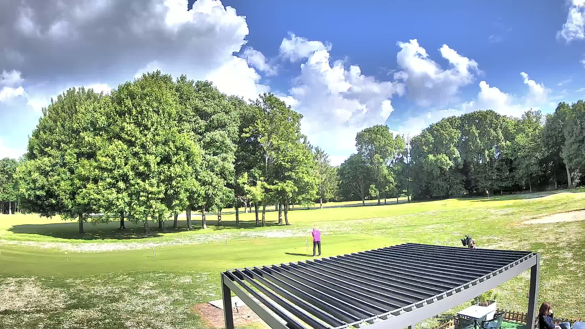 A golfer tees off on a partly sunny day on a green golf course surrounded by lush green trees.