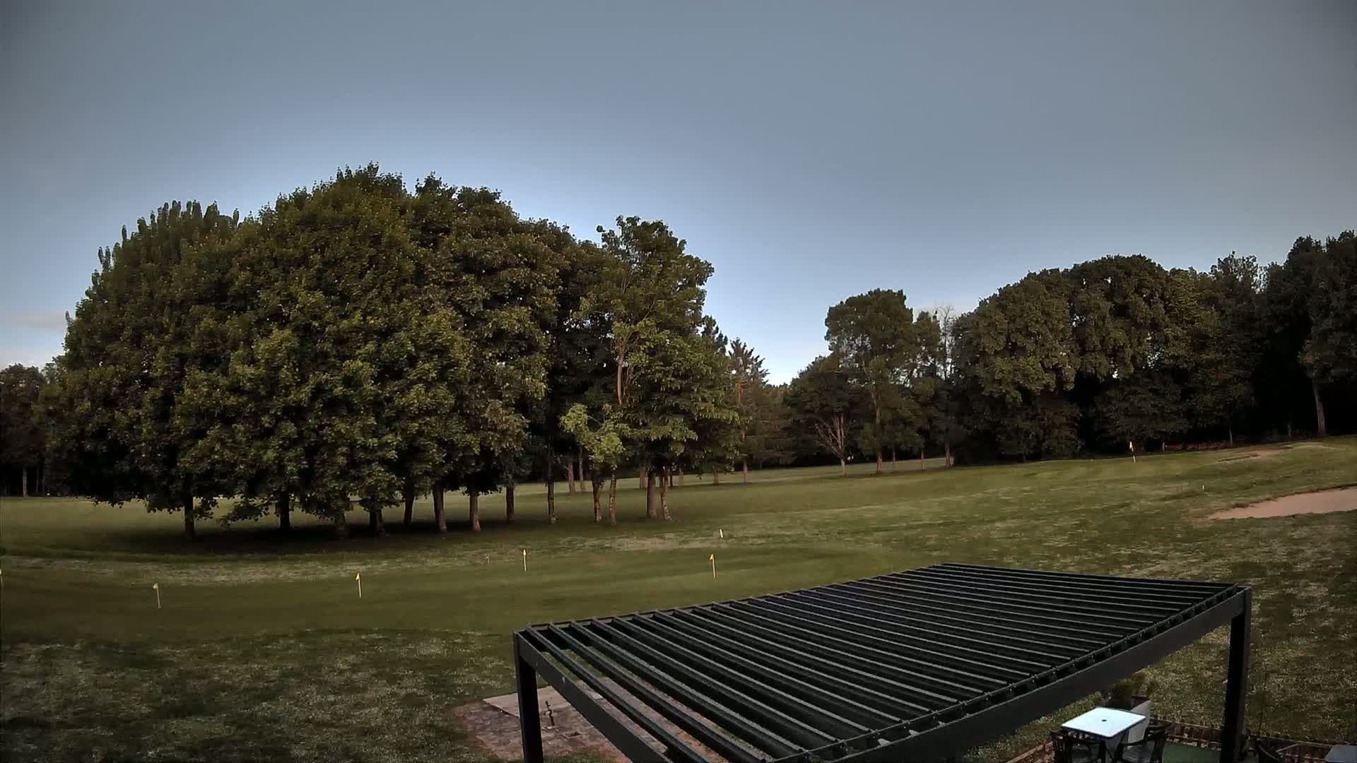 A mostly clear sky overlooks a grassy putting green surrounded by lush green trees, with a dark pergola in the foreground.