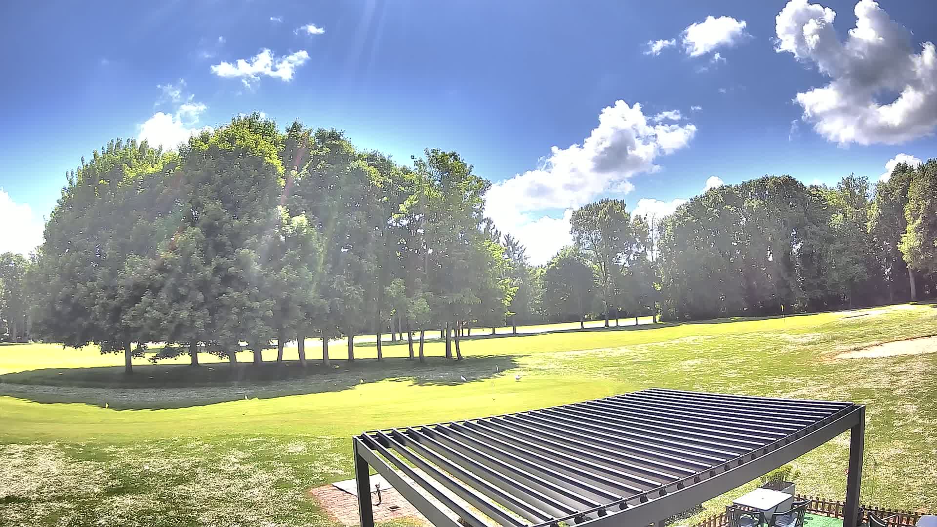 A sunny day on a golf course, with a mostly clear blue sky and fluffy white clouds, shows a green lawn dotted with trees and a modern pergola in the foreground.