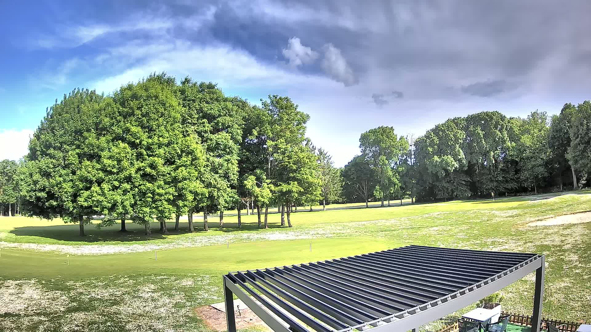 A partly cloudy sky overlooks a green golf course dotted with deciduous trees and a modern pergola.