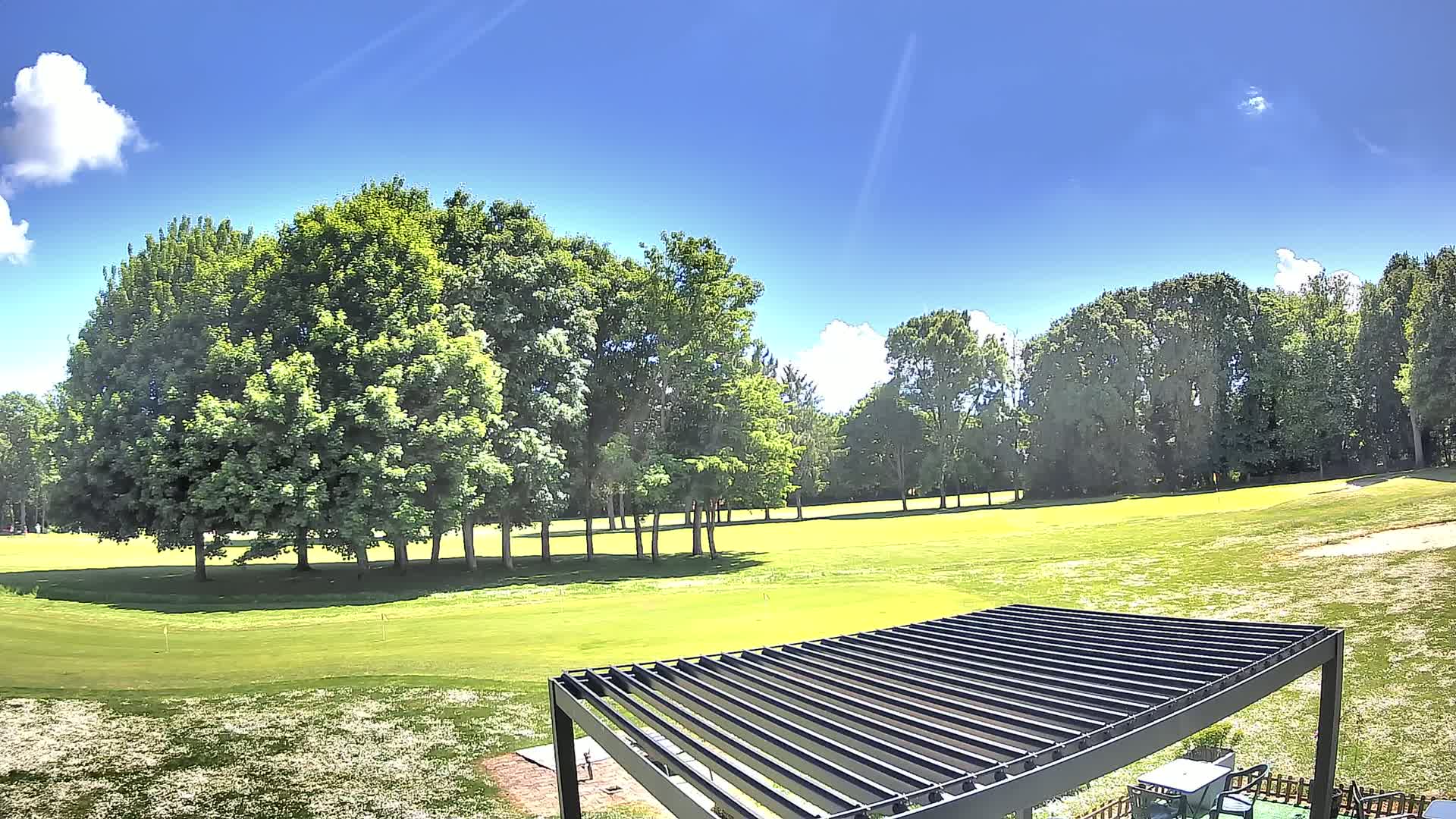 A sunny day reveals a golf course with lush green grass, bordered by a line of full, leafy trees under a mostly clear blue sky.