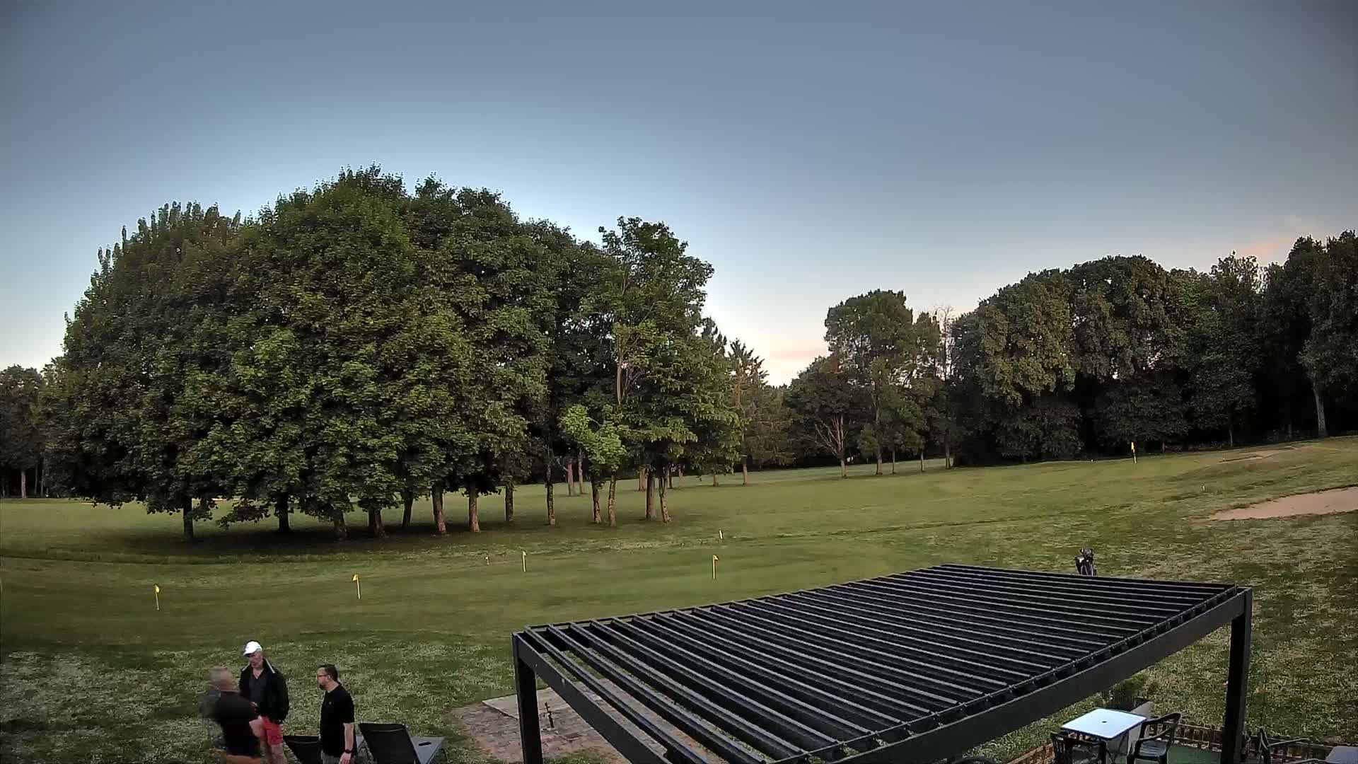 A grassy golf course with several trees in the background under a clear, twilight sky, and three men standing near a pergola in the foreground.