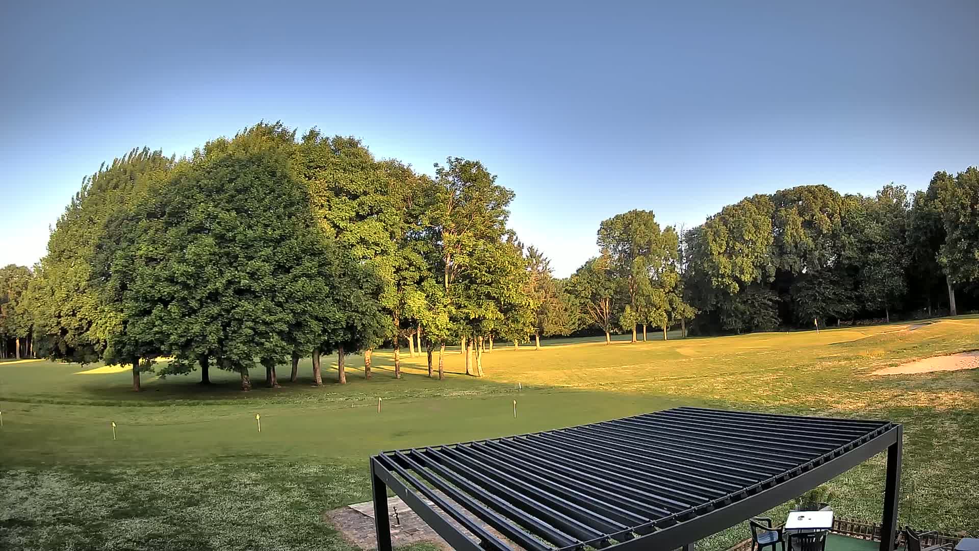 A golf course with lush green grass and many leafy trees is visible under a clear blue sky, partially obscured by a dark slatted pergola in the foreground.