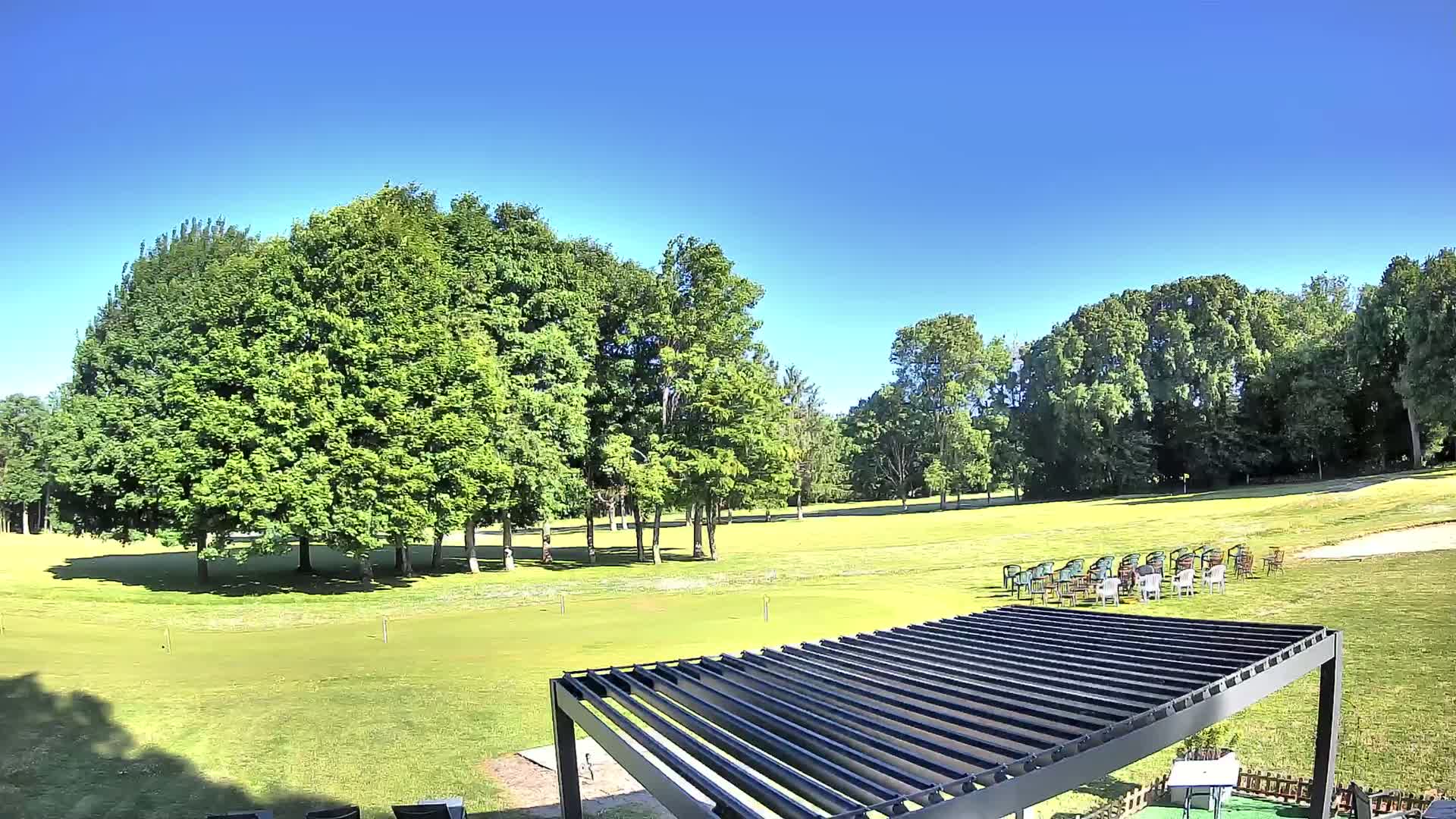 A grassy area with many green trees under a clear blue sky, featuring a modern pergola and a cluster of chairs.