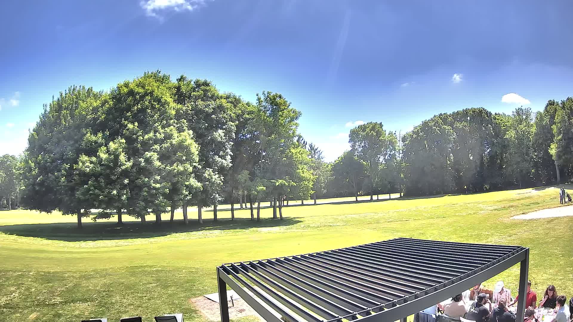 A group of people are seated under a pergola on a sunny day overlooking a grassy golf course surrounded by lush green trees.