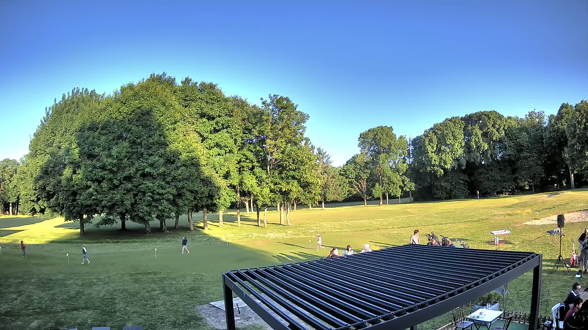 A putting green surrounded by lush green trees under a clear blue sky, with several people playing and others gathered near a modern pergola.