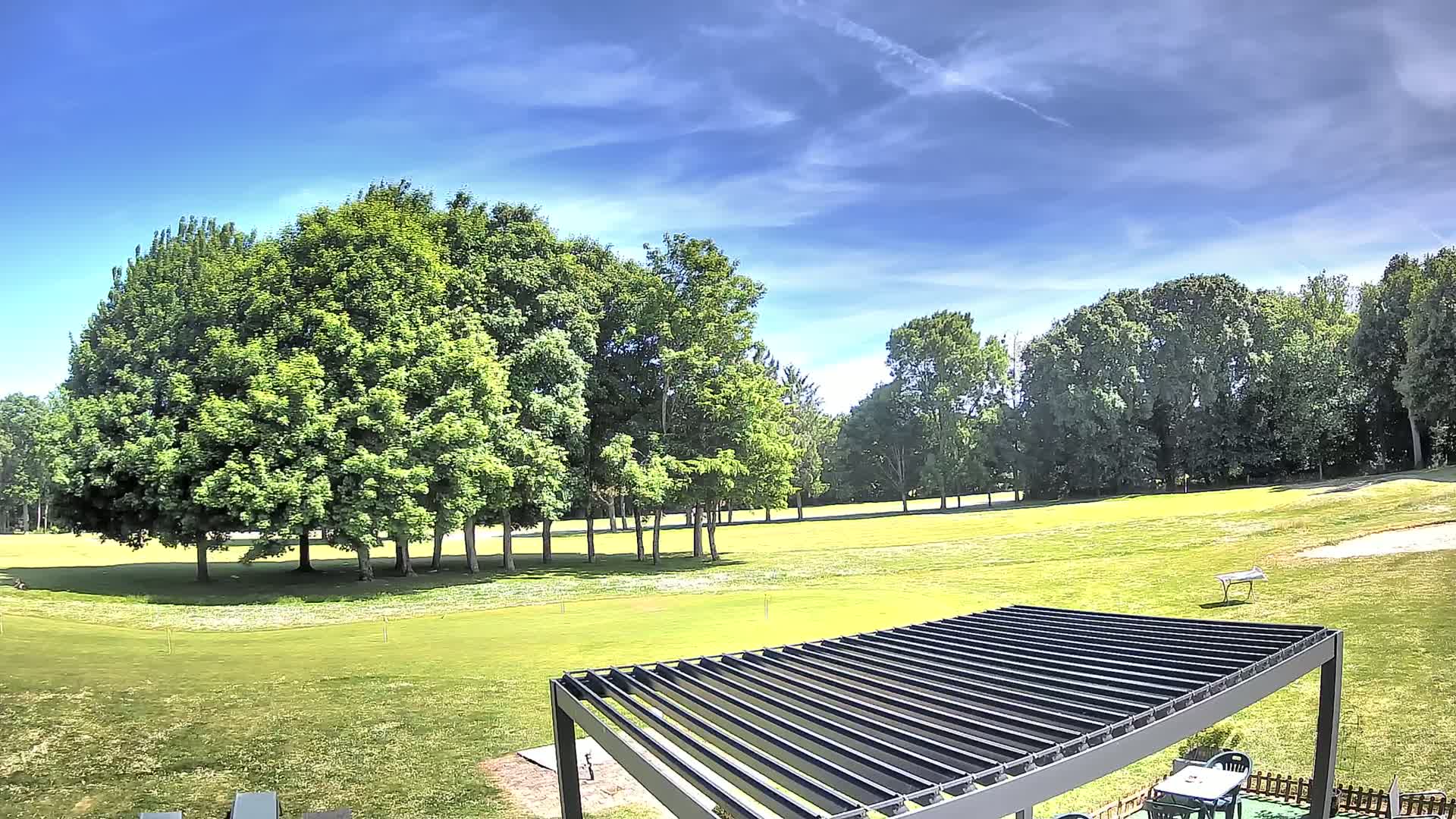 A partially visible black pergola sits in the foreground of a grassy area overlooking a golf course under a mostly sunny sky.