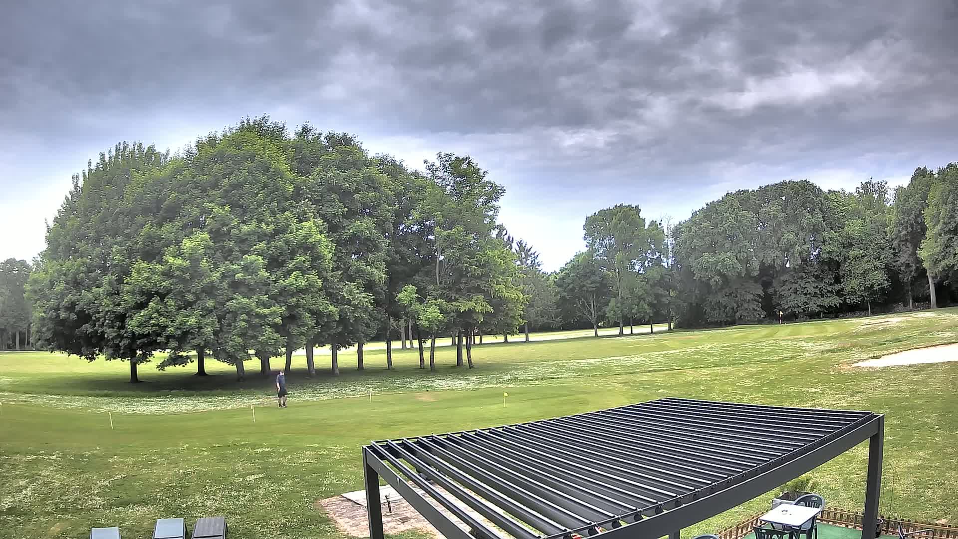 A person stands on a green golf course under a cloudy sky, near a modern pergola and lounge chairs.