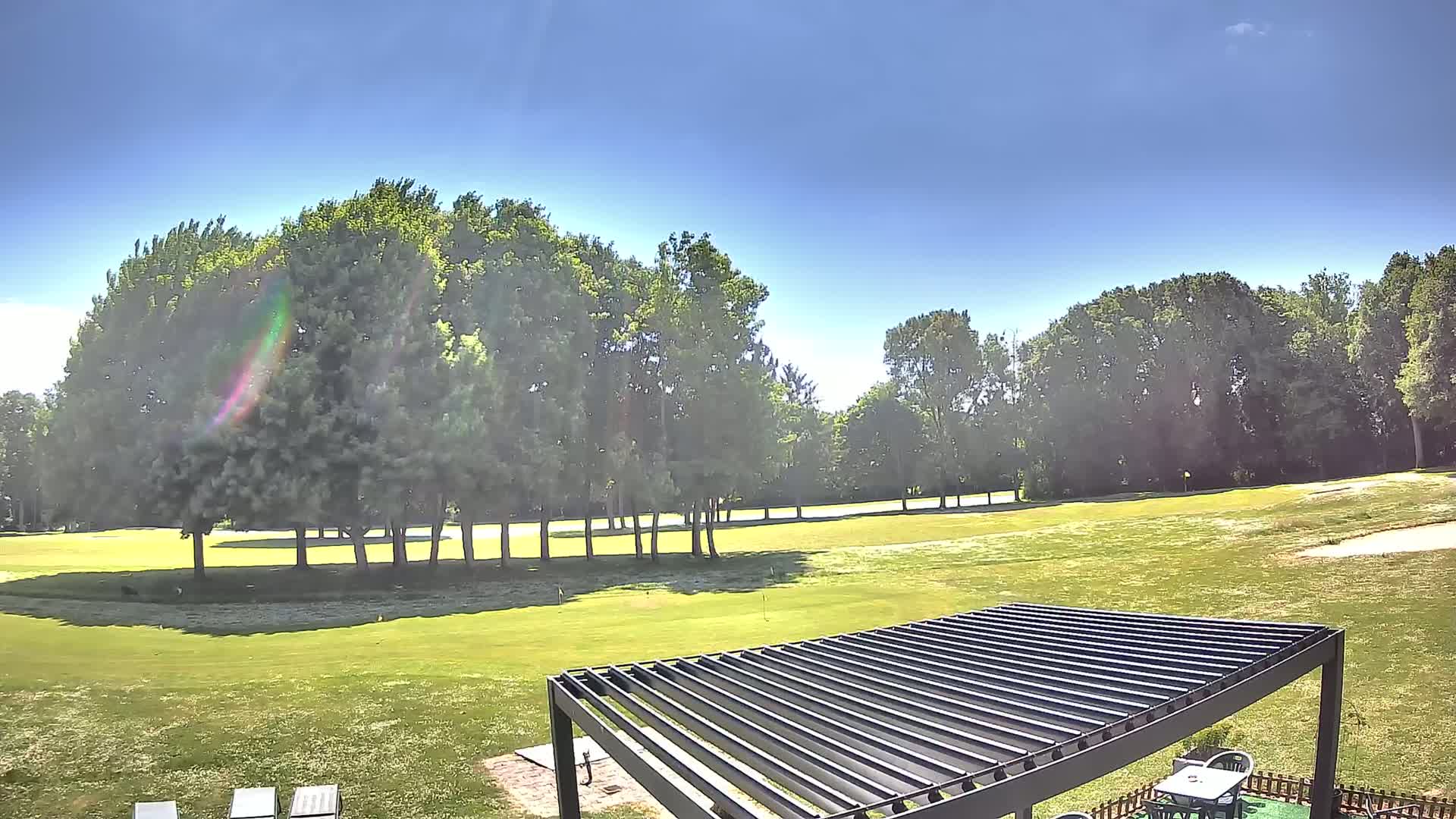 A sunny day reveals a golf course with lush green grass, bordered by tall, leafy trees, and a dark-colored pergola partially visible in the foreground.