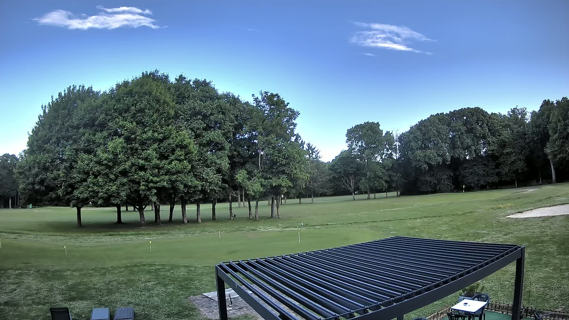 A golf course with green fairways and sand traps is visible beyond a black pergola on a partly sunny day.