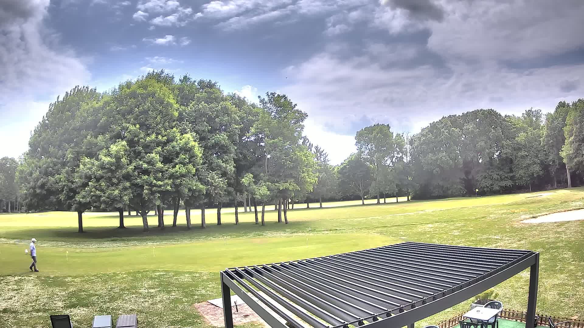 A golfer walks across a green golf course under a partly cloudy sky, near a modern pergola and patio furniture.