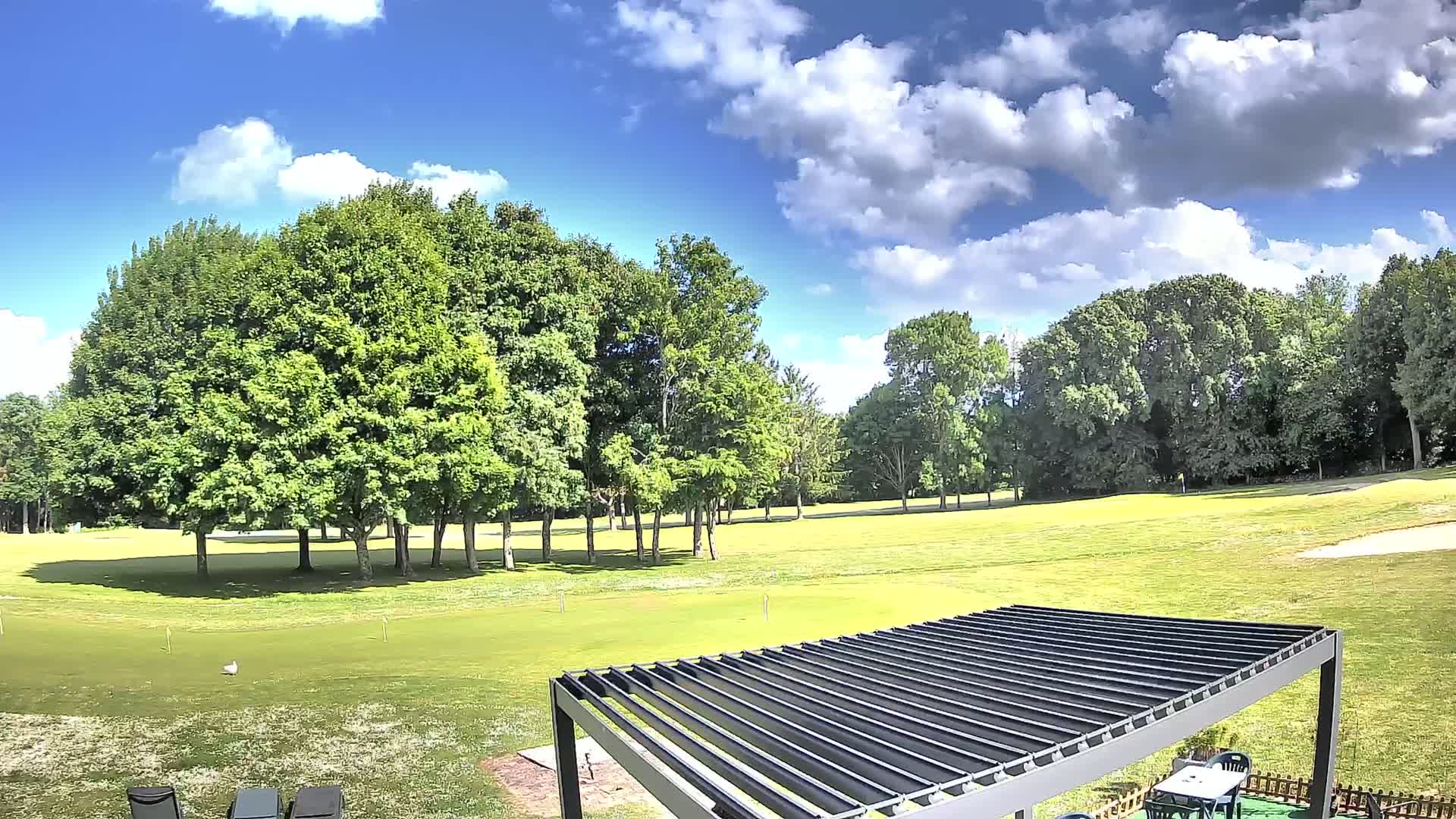 A mostly sunny day with fluffy white clouds shows a green golf course surrounded by lush green trees and a modern pergola with a table and chairs.
