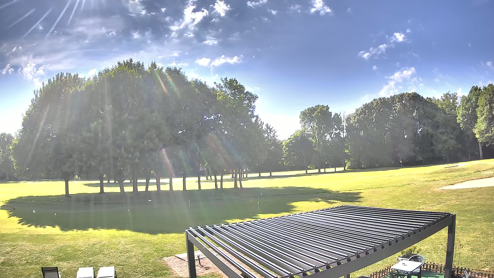 A sunny day with partly cloudy skies shows a putting green surrounded by lush green trees and a slatted pergola in the foreground.