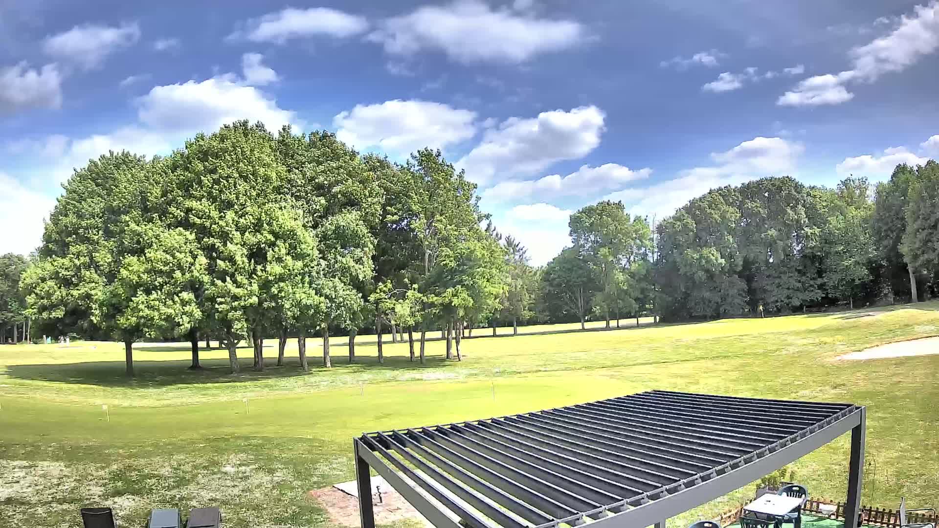 A partly cloudy sunny day reveals a sprawling green golf course, bordered by lush green trees, and a modern pergola in the foreground.