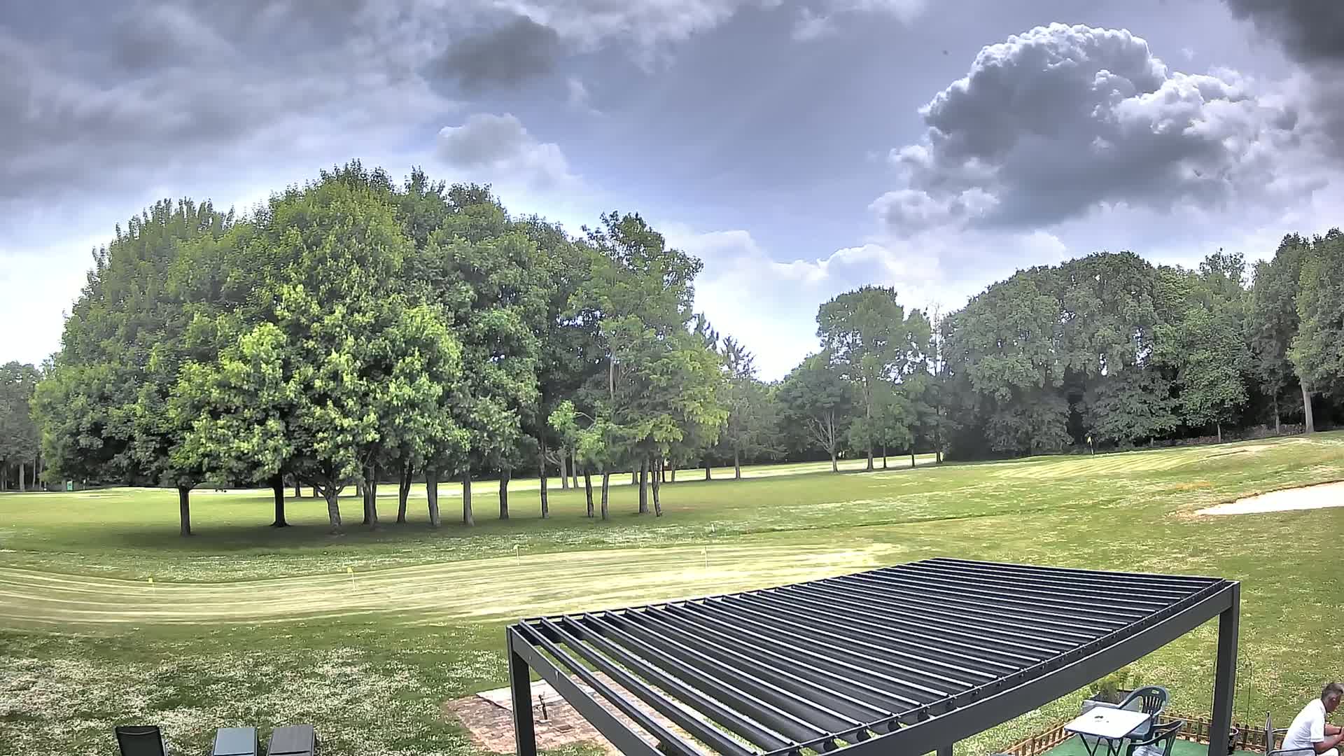 A mostly sunny day with some clouds shows a green golf course surrounded by lush trees, and a black pergola with a table and chairs sits in the foreground.