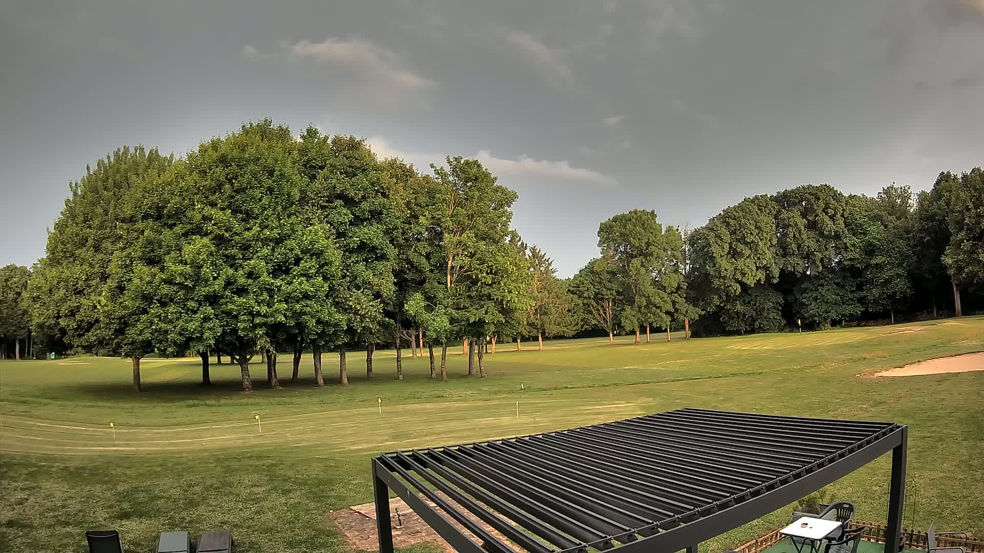 A golf course with green grass, numerous full, leafy trees, and a dark-colored pergola is visible under a mostly cloudy sky.