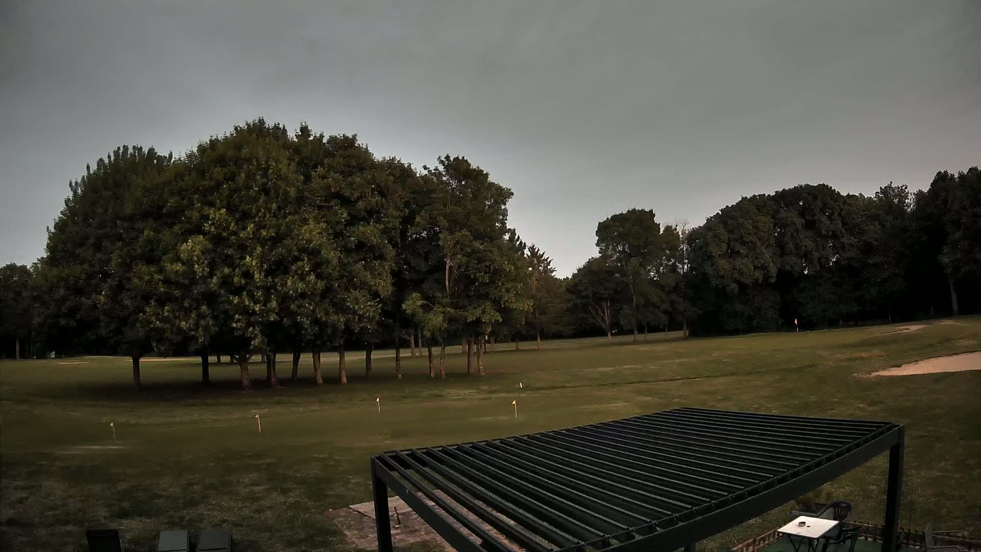 A mostly overcast sky hangs over a putting green surrounded by trees and a dark metal pergola.