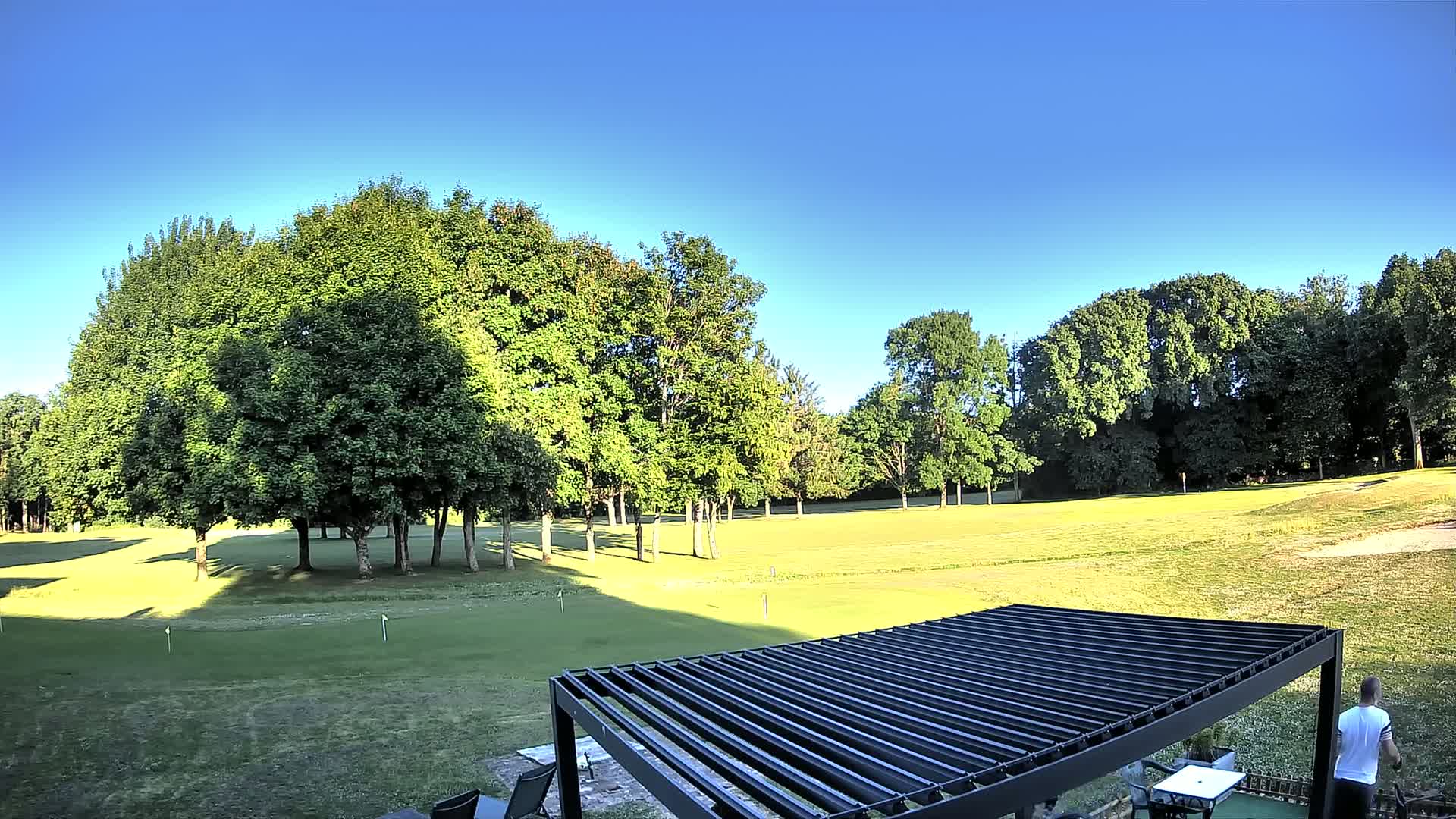 A sunny day reveals a golf course with lush green grass surrounded by tall, leafy trees, partially obscured by a black pergola.