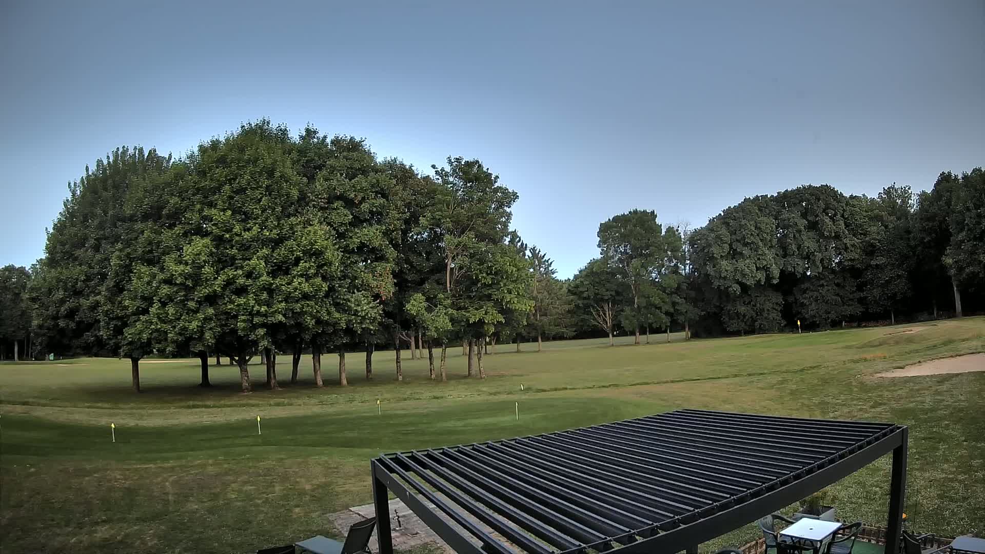 A grassy golf course with numerous green trees under a clear blue sky is partially visible from behind a dark-colored pergola.