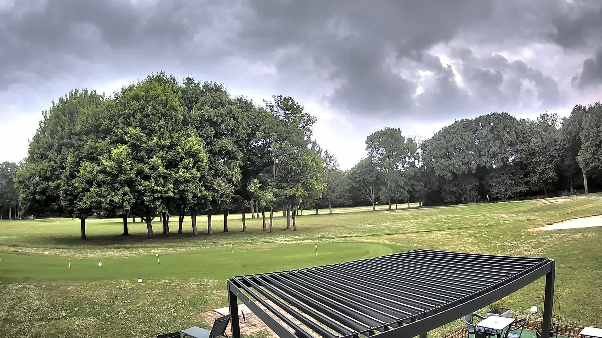 A golf course with lush green fairways and putting greens, bordered by a line of tall trees, is viewed from under a dark gray, overcast sky.