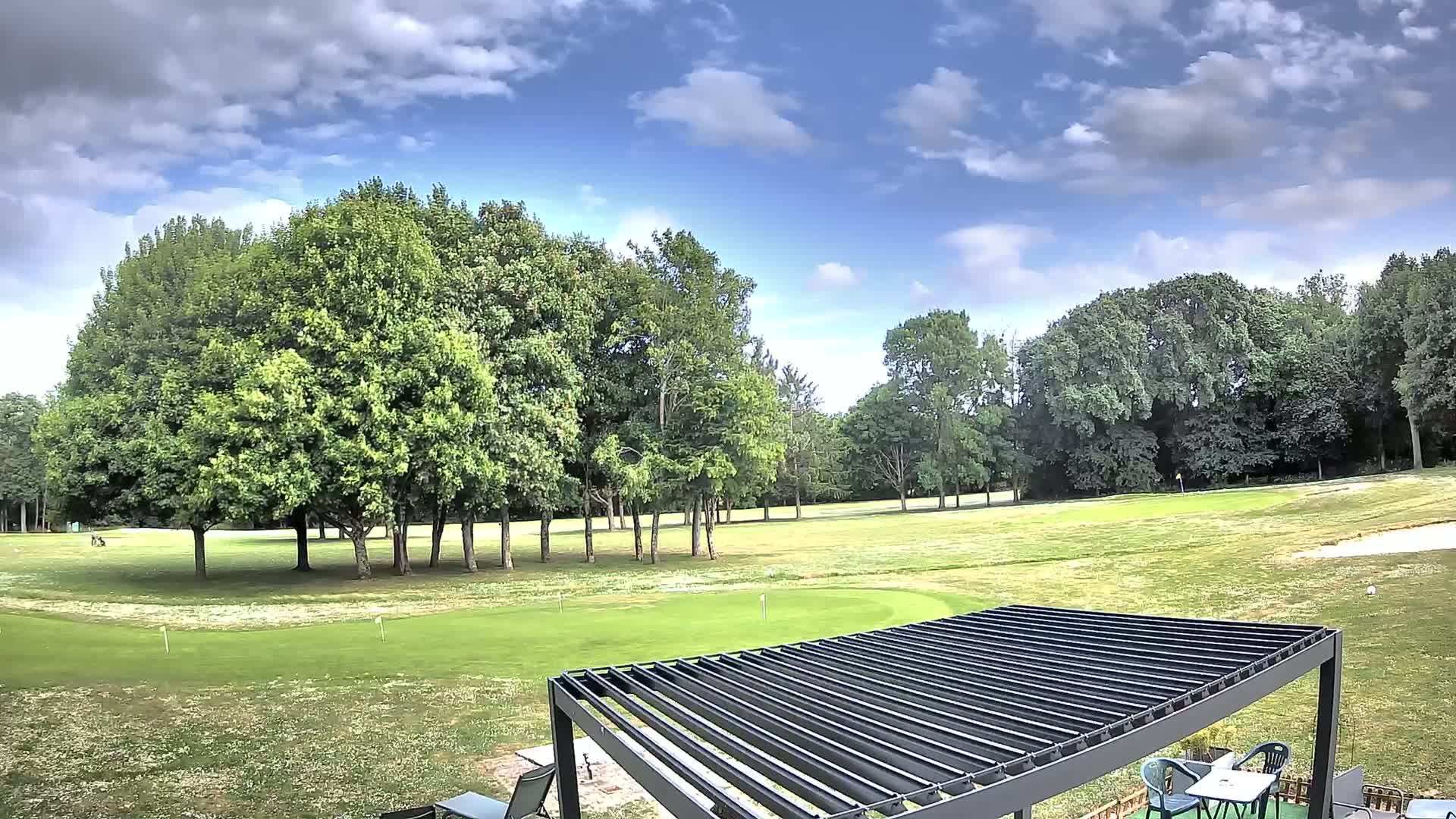 A partly cloudy sky overlooks a golf course with lush green grass, several full, green trees, and a modern pergola with outdoor furniture.