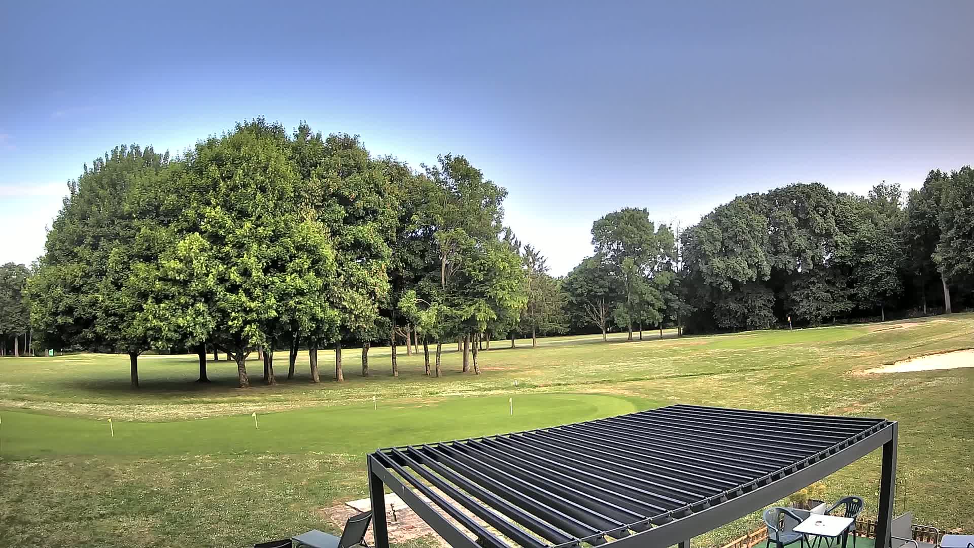 A putting green bordered by lush green trees under a clear blue sky is partially visible beneath a dark pergola.