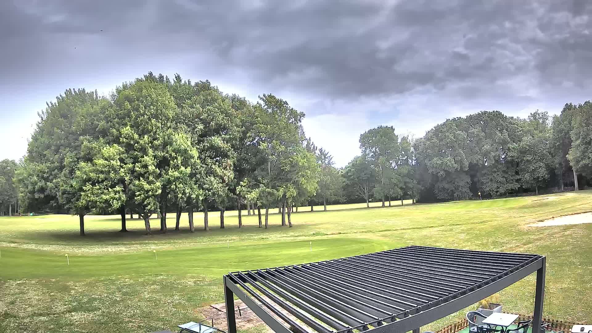 A golf course with a dark-colored pergola in the foreground, under a cloudy sky.