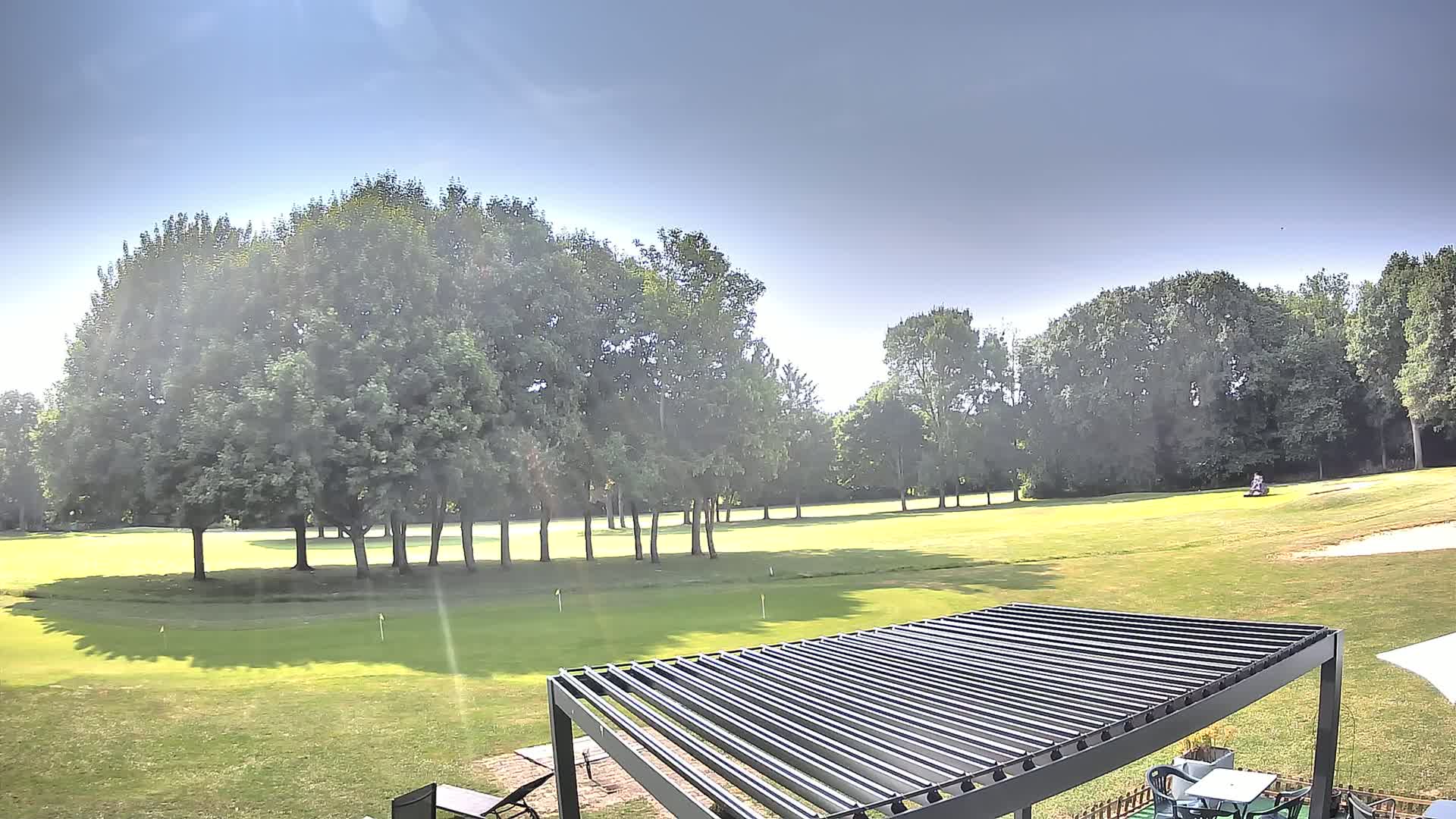A sunny day on a golf course shows a green fairway, several trees, and a slatted pergola in the foreground.