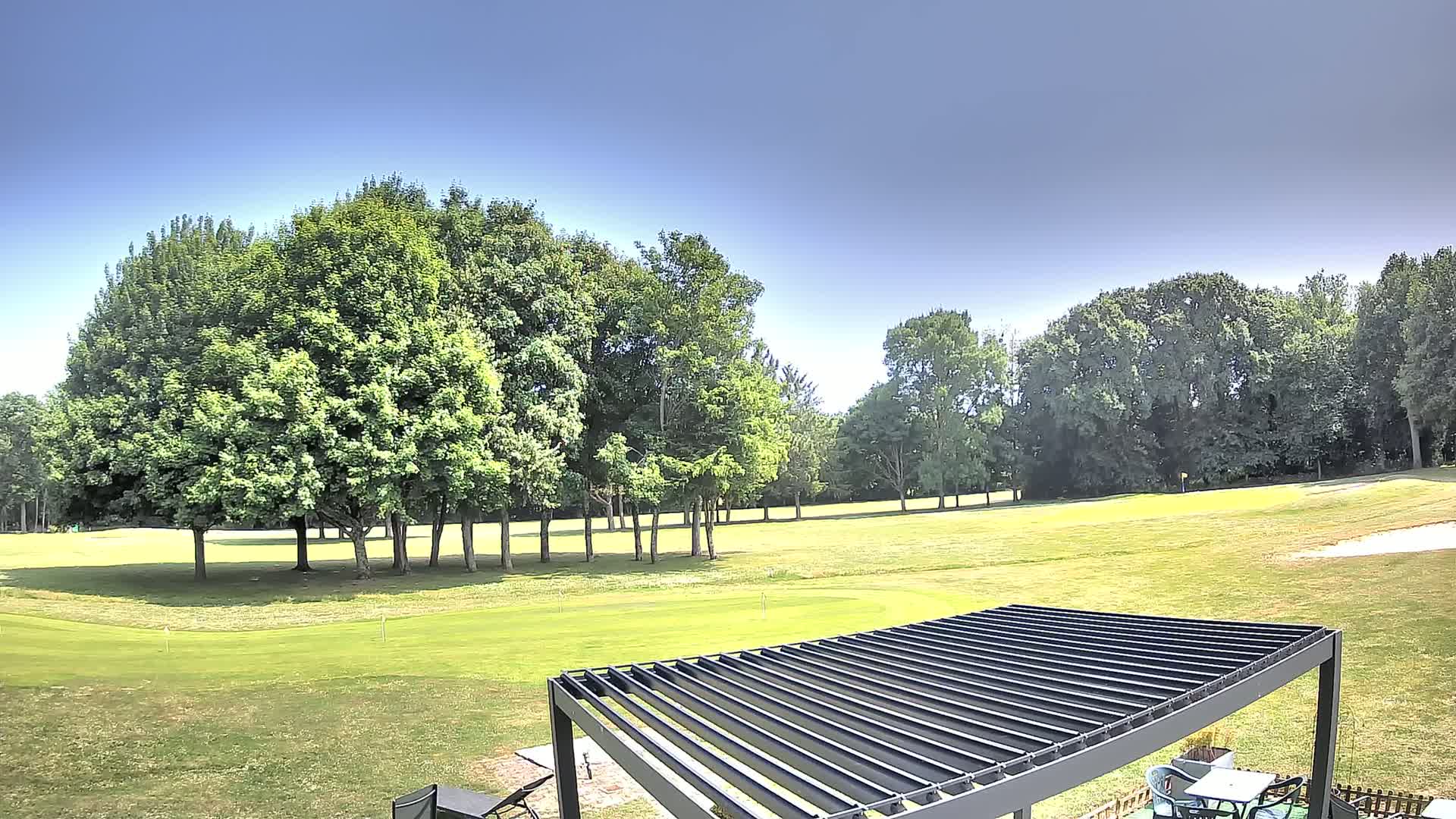 A golf course with lush green grass and many full, green trees under a clear, sunny sky is partially visible from under a dark gray pergola.