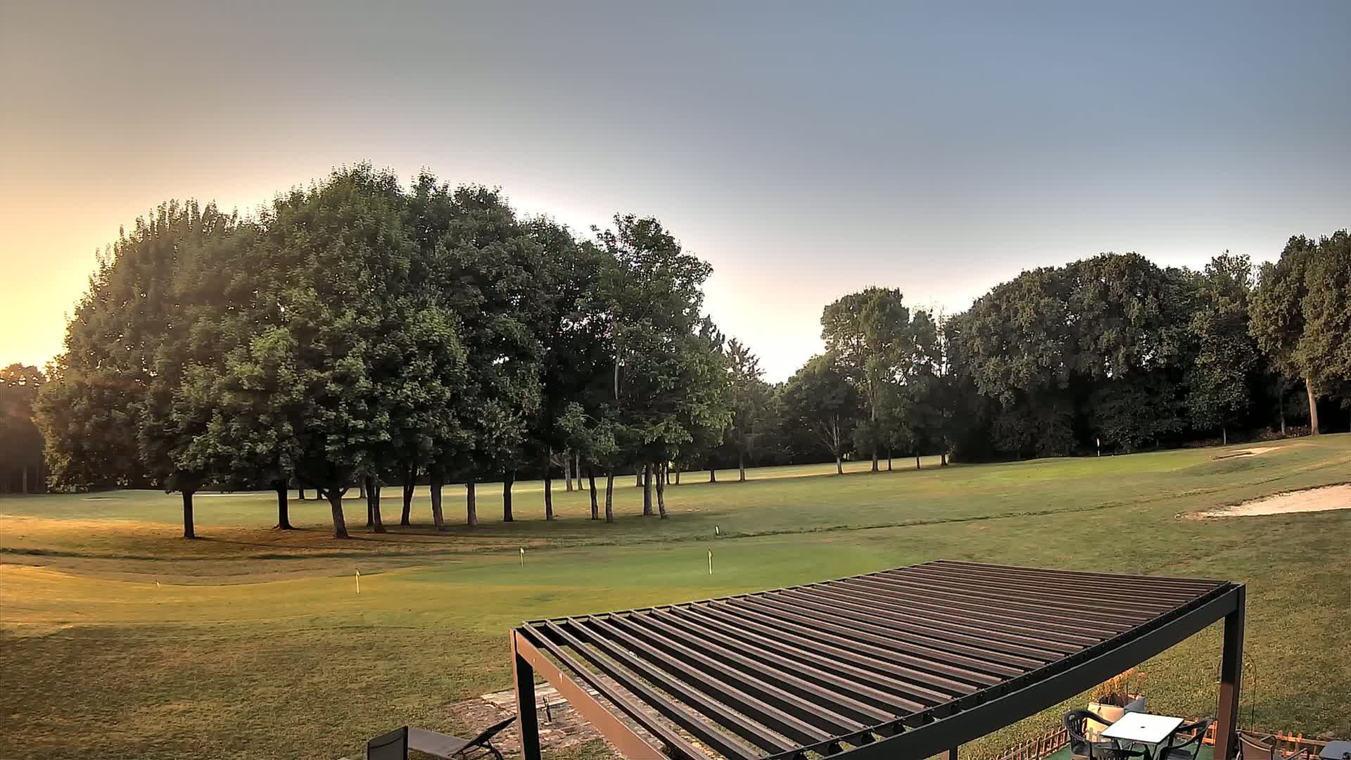 A sunlit golf course with a cluster of trees in the mid-ground is viewed from a patio with a slatted pergola.