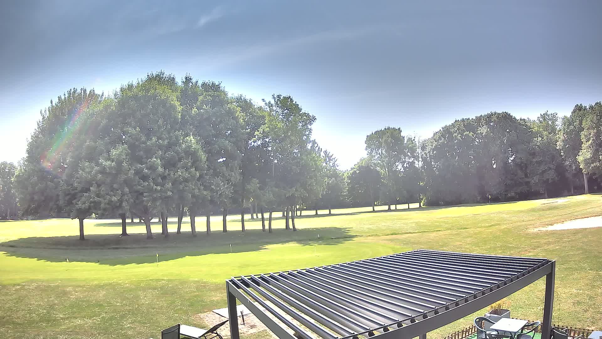 A sunny day reveals a lush green golf course bordered by tall trees, partially visible behind a modern metal pergola.