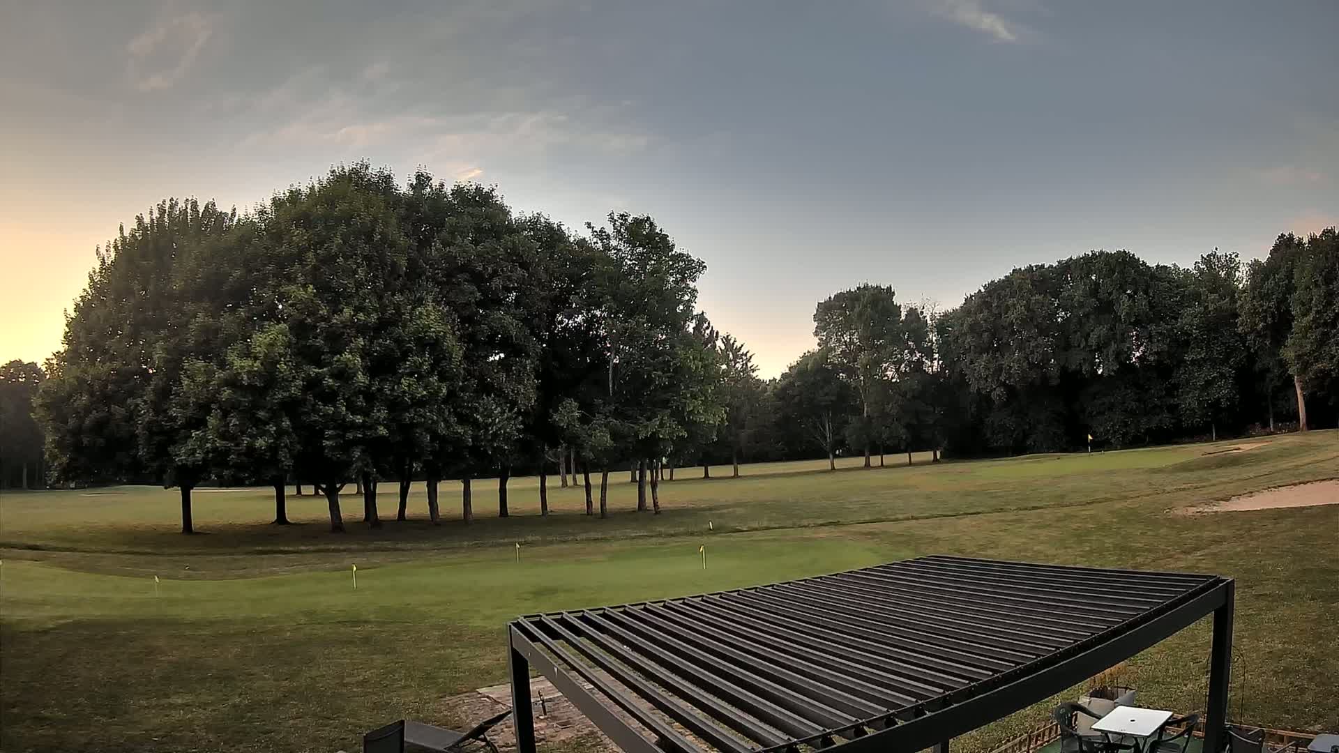 A grassy golf course, bordered by lush green trees, is visible under a partly cloudy, dusky sky, with a dark metal pergola in the foreground.
