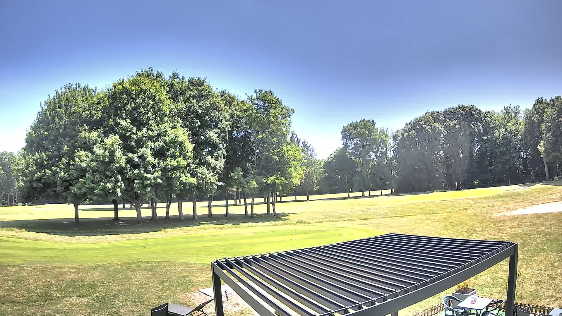 A green golf course with a cluster of trees in the background under a clear, sunny sky, partially obscured by a dark-colored pergola in the foreground.