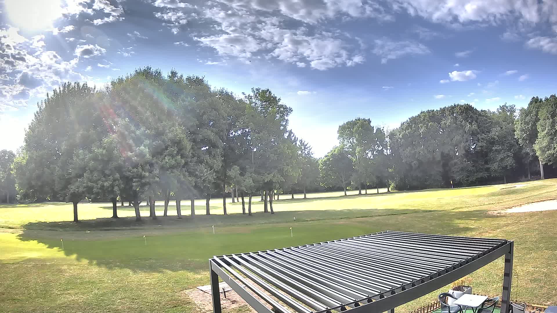 A sunny day with partly cloudy skies shows a view of a putting green surrounded by lush green trees and a slatted metal pergola in the foreground.