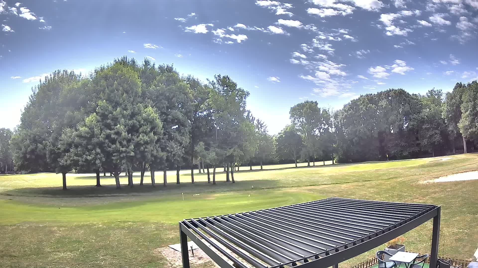A partly sunny day reveals a golf course with green grass and trees, partially obscured by a dark metal pergola.