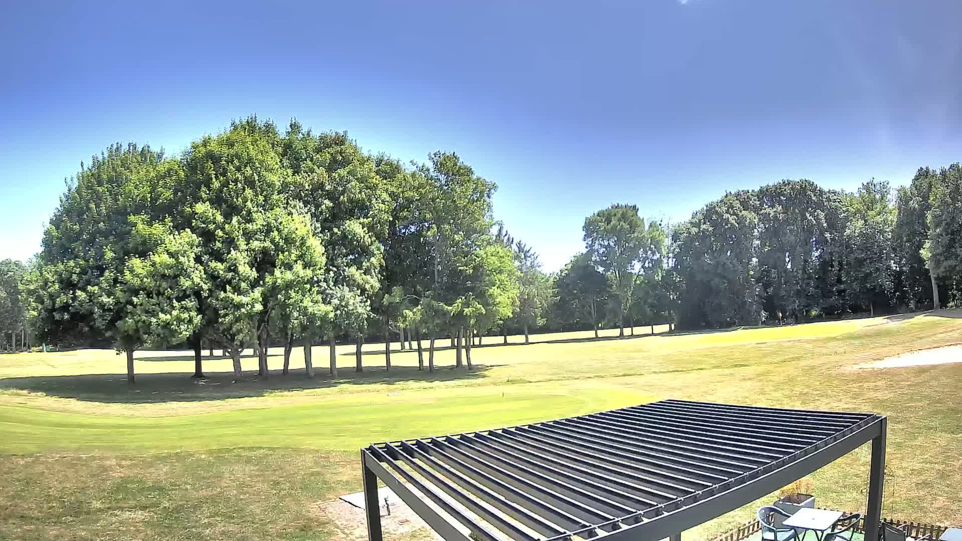 A sunny day on a golf course, featuring a grassy fairway, several green trees, and a dark metal pergola in the foreground.