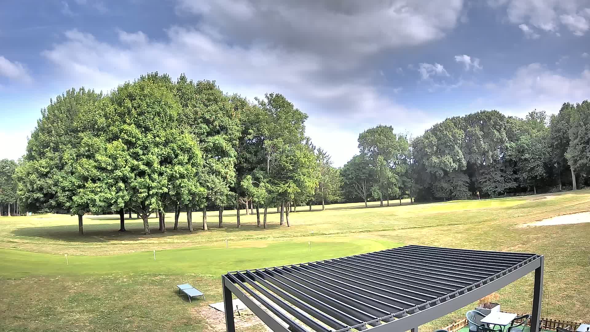 A partly cloudy sky overlooks a golf course with lush green grass, a cluster of leafy trees, and a dark gray pergola.