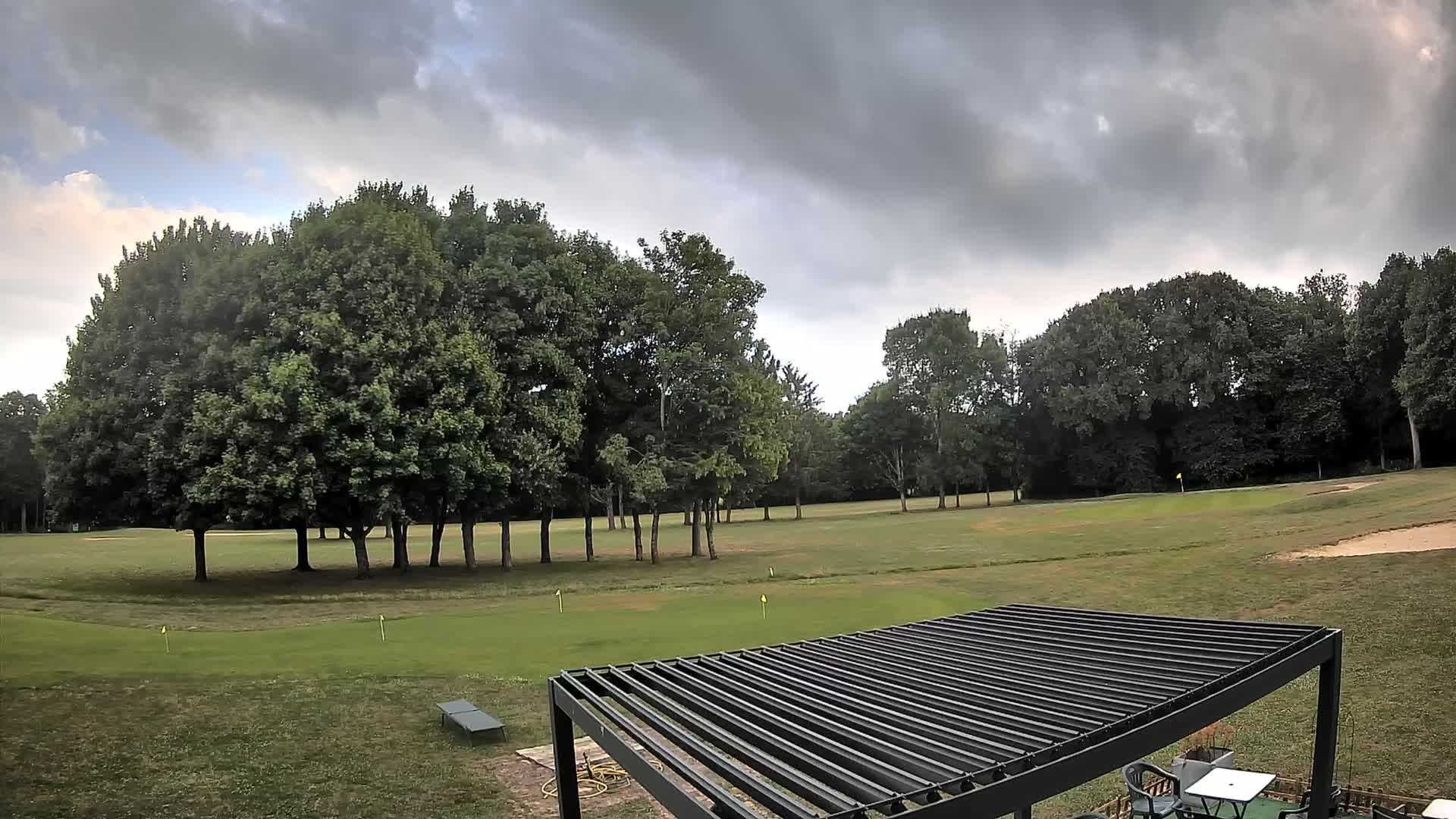 A partly cloudy sky overlooks a golf course with lush green grass, a cluster of leafy trees, and a dark gray pergola.