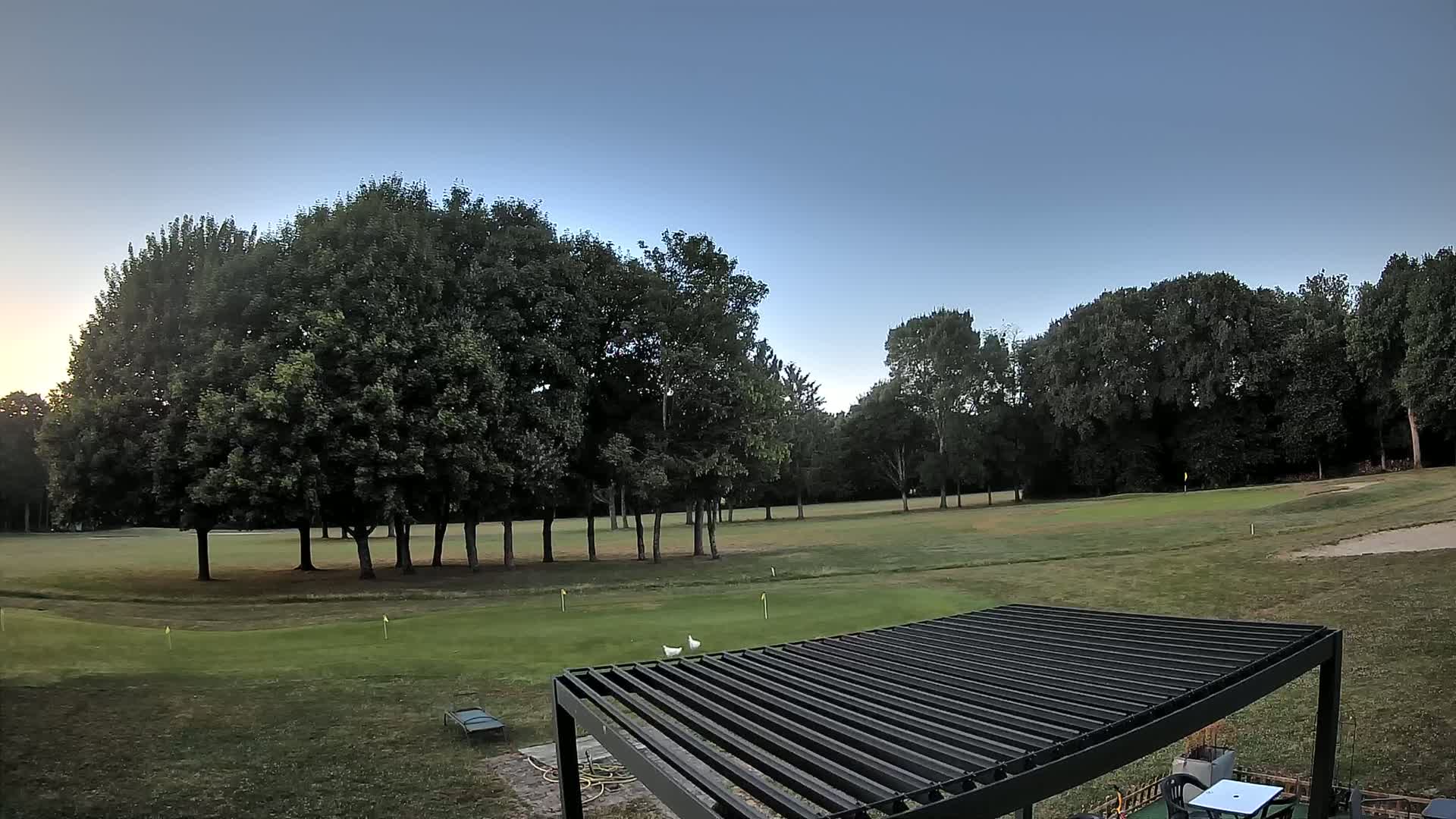 A golf course with a cluster of trees in the background under a clear, twilight sky, partially obscured by a dark-colored pergola.