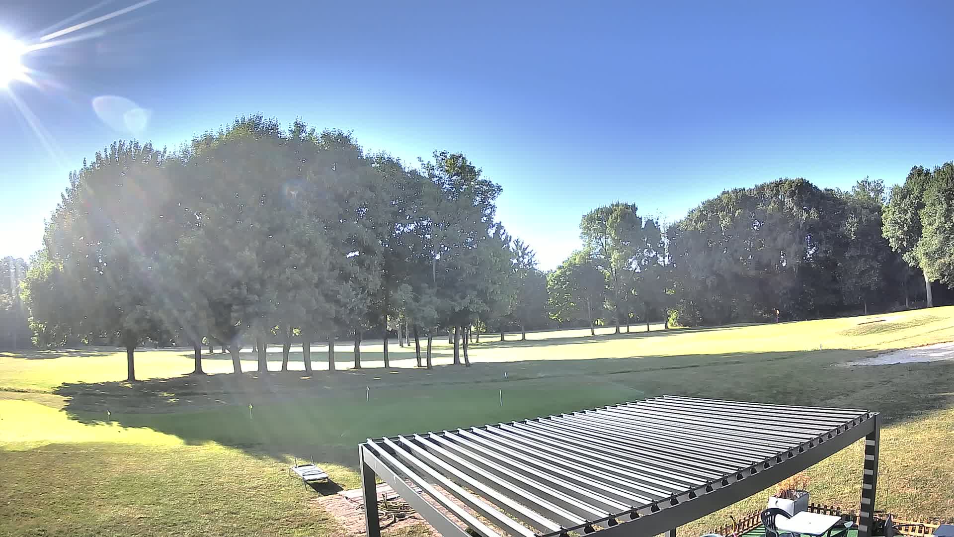 A sunny day reveals a putting green bordered by trees, partially obscured by a metal pergola in the foreground.
