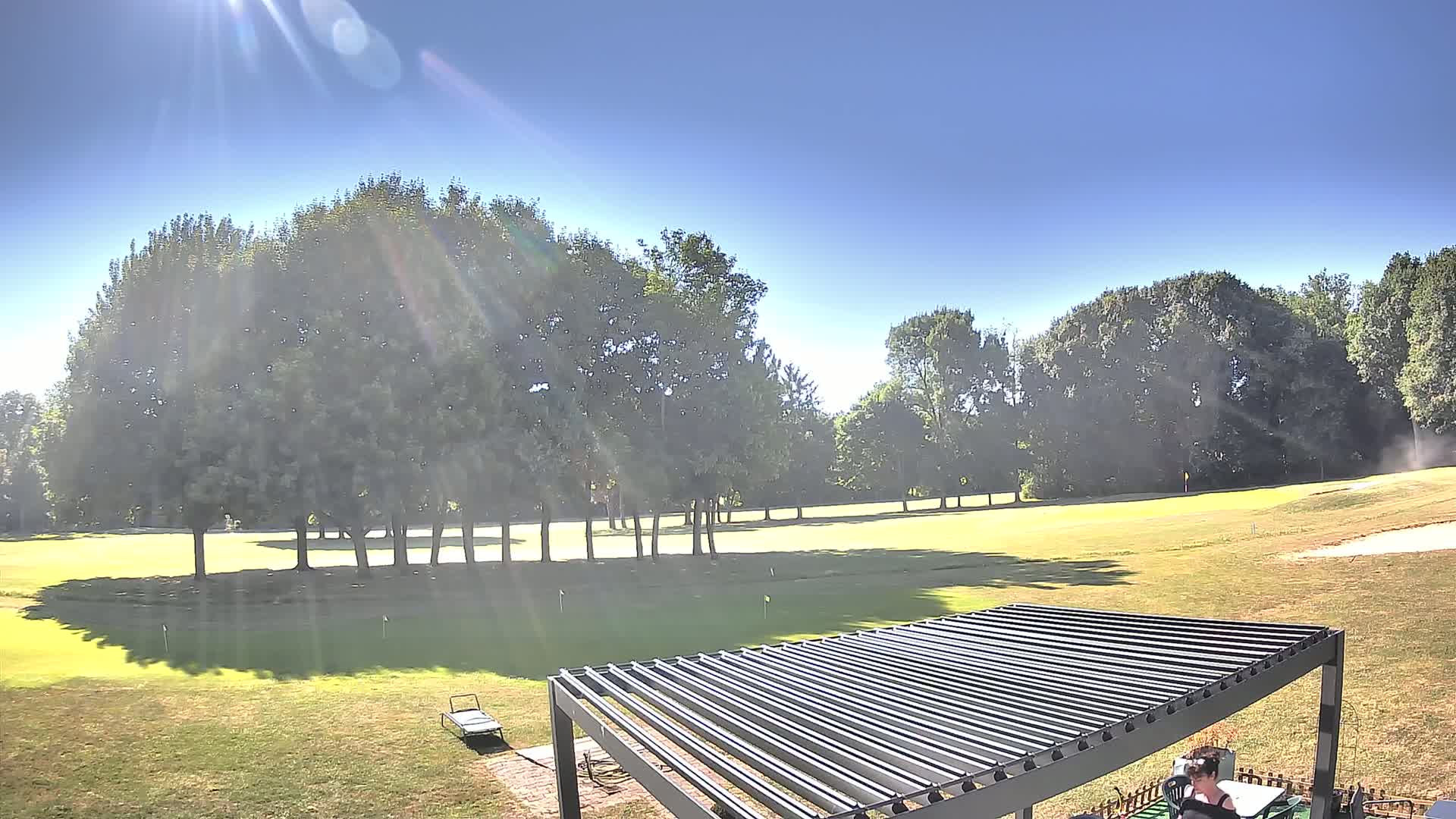 A sunny day reveals a golf course framed by leafy green trees, partially visible behind a modern slatted pergola.