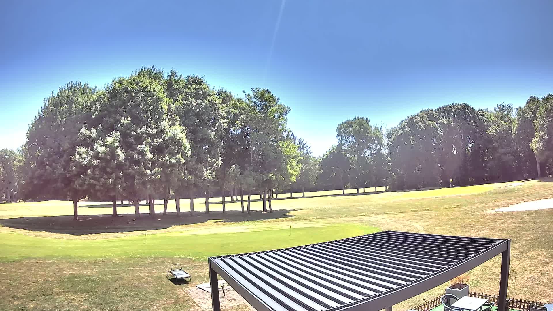 A black pergola sits on a grassy area overlooking a golf course on a sunny day.