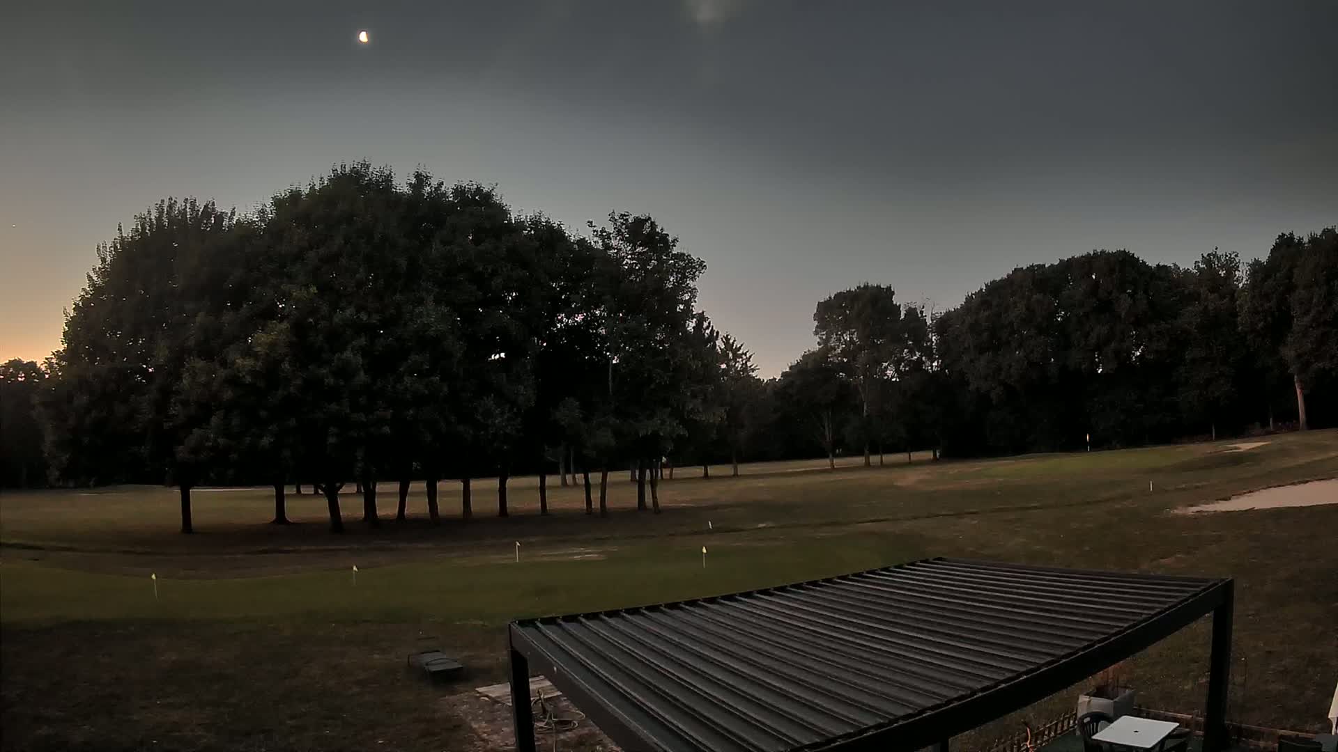 A crescent moon hangs in the twilight sky above a mostly-treed golf course, partially visible from behind a dark pergola.