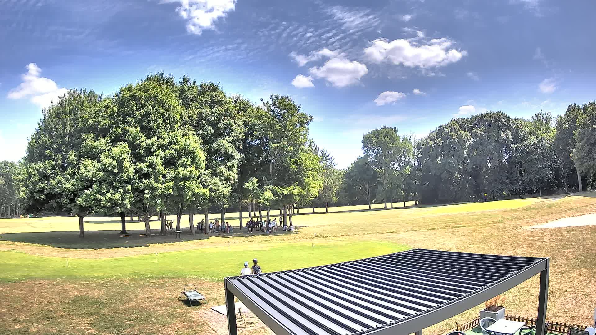 A sunny day on a golf course shows a group of people gathered under trees, with a modern pergola in the foreground.