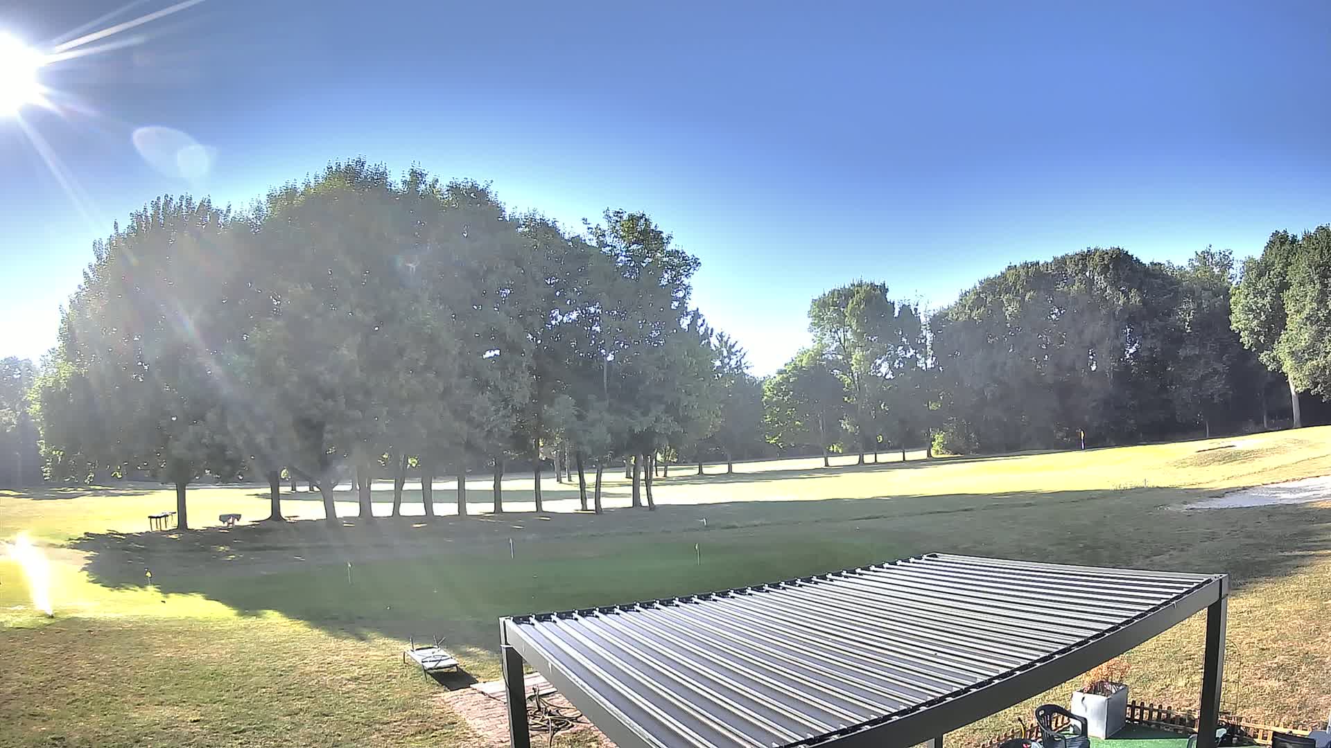 A sunny day reveals a golf course with a line of trees in the background, and a pergola in the foreground.