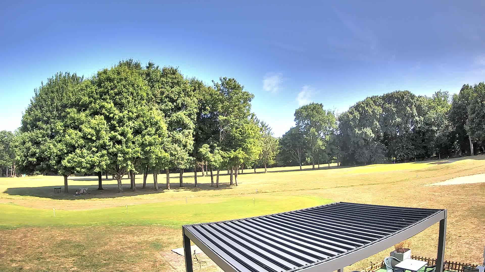 A golf course with a mostly sunny sky is viewed from a patio with a dark-colored pergola.