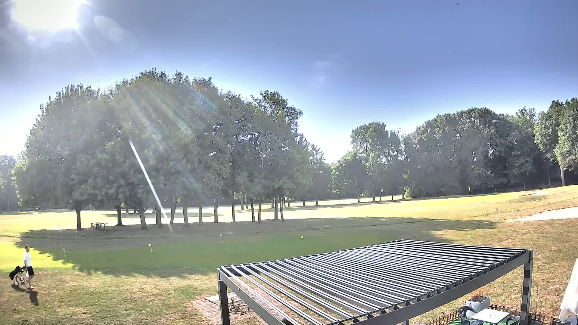 A golfer walks across a sunny golf course surrounded by trees and a modern pergola is in the foreground.